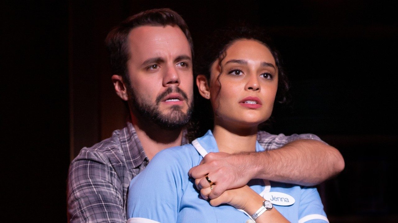 Shawn W. Smith, a white man with dark brown hair, beard, and moustache, stands bahind Jisel Soleil Ayon, a Black woman with dark curly hair pulled into a ponytail. She wears a powder blue diner uniform. He wears a grey plaid shirt and a wedding ring. He puts one arm around her from behind, holding her to him.
