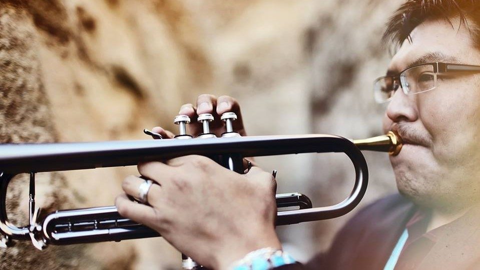  Trumpeter Delbert Anderson, an Indigenous man with dark short hair and glasses wearing a turquoise necklace and bracelet, plays his trumpet in front of rocky walls.