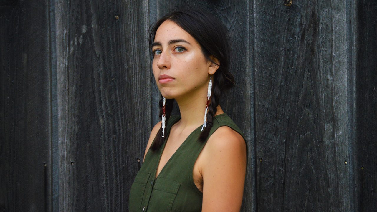  Mali Obomsawin is an Indigenous woman with hazel eyes and dark shoulder length hair parted down the middle and braided into pigtails, wears an olive green dress and traditional white beaded earrings.