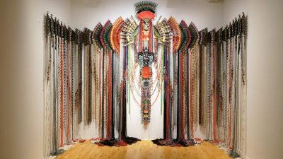  A work by Malaysian fiber artist Anne Samat hangs on a gallery wall, parts of the piece trailing on the floor in a specific pattern.