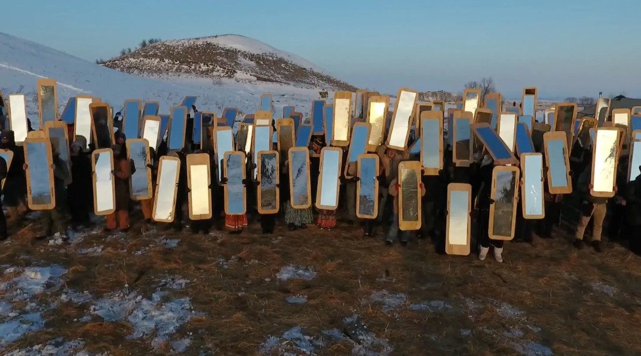  Cannupa Hanska Luger's "Mirror Shield Project," 2016. A large group of people stand in a field somewhere that snow and ice is melting, a snow-covered hill and field visible behind them. They all wear cold-weather attire and hold floor-length mirrors fixed to natural wood backers and point the mirrors towards the sun, away from the melting snow.