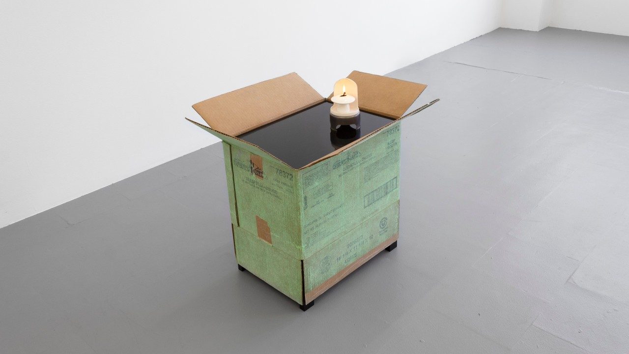  Peggy Chiang's "Invitation to travel," 2022. A green cardboard box sits on a grey floor, the top flaps open. A black, shiny, plexiglass material fits just inside the opening. On the plexiglass sits a small, white, burning tealight.