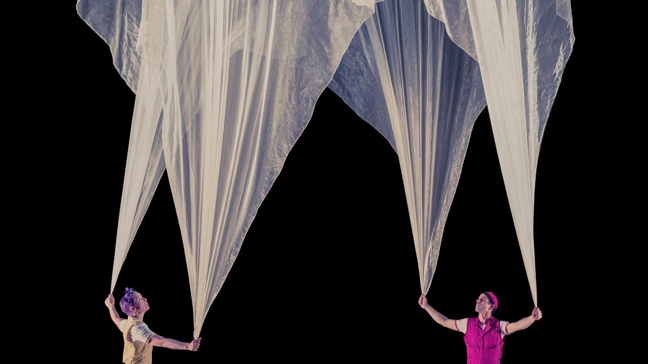  The cast members of Acrobuffos, a woman with purple hair and a yellow jumpsuit and a man with a red beanie and a red jumpsuit, each hold two corners of an enormous piece of white chiffon. The fabric balloons upwards in front of a black background, caught by the air from fans.