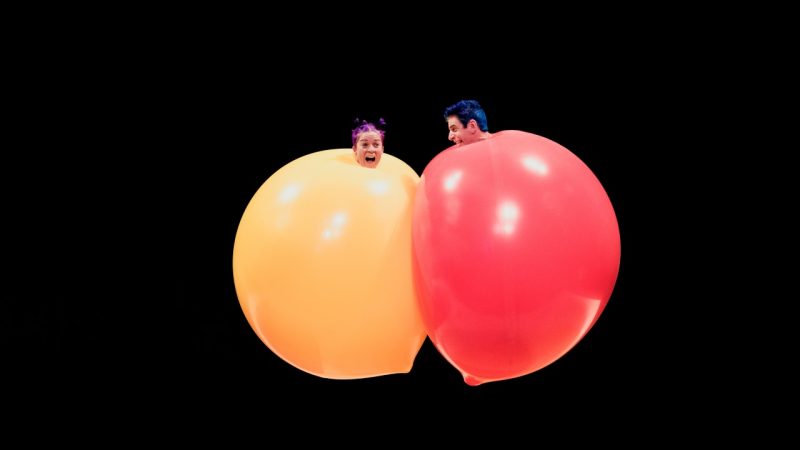  The cast members of Acrobuffos jump into the air in front of a black background and bounce into each other, fully engulfed in large yellow and red balloons, apart from their heads.
