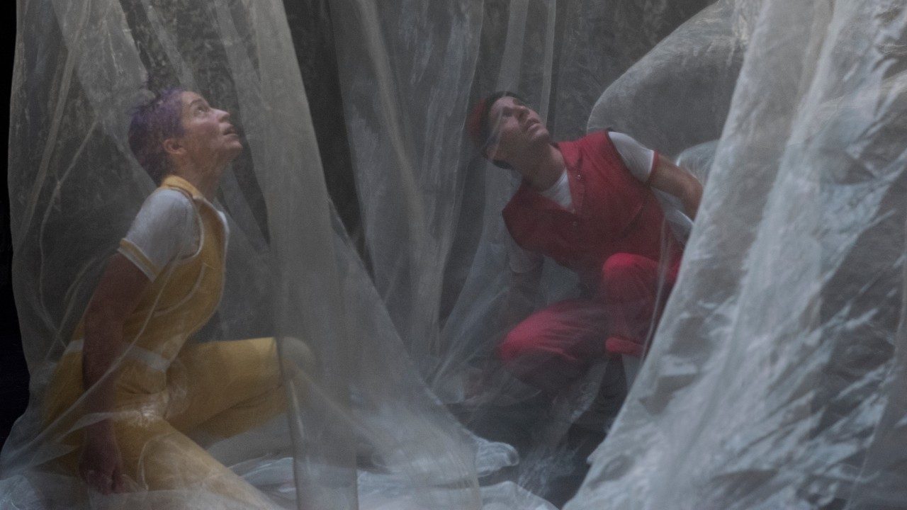  The cast members of Acrobuffos, a woman with purple hair and a yellow jumpsuit and a man with a red jumpsuit, crouch under off-white chiffon fabric and look upwards.