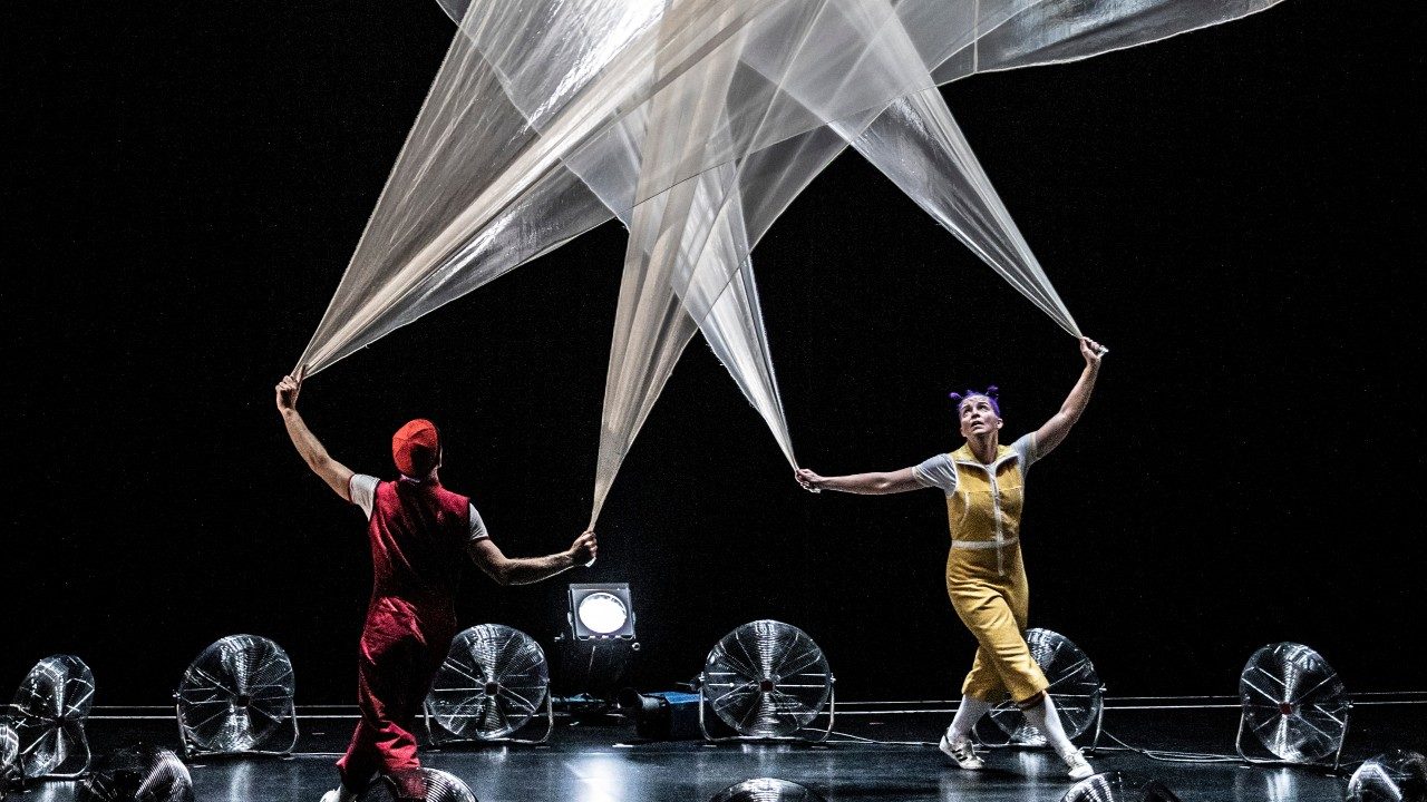  Two cast members of Acrobuffos stand in middle of a circle of industrial fans. They each hold onto two corners of a large piece of thin white fabric. The cast members walk in circles as the fans catch the fabric and make it float up into the air.