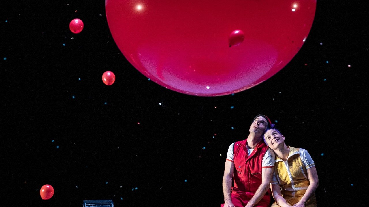  Two cast members of Acrobuffos sit together on stage, a man in a red jumpsuit on the left, and woman in a yellow jumpsuit on the right, leaning her head on the man's shoulder. They both gaze upward at a large red balloon and several smaller red balloons, all floating in the air above them. Behind them is a starry night sky.