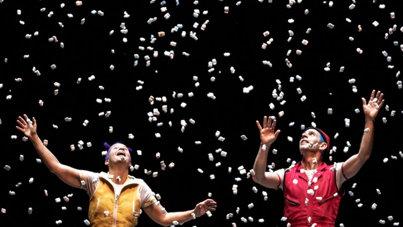  Two cast members of Acrobuffos stand beneath packing peanuts that look like snow as a large quantity of it drops onto them from above. The woman on the left wears a yellow jumpsuit, and the man on the right wears a red one.