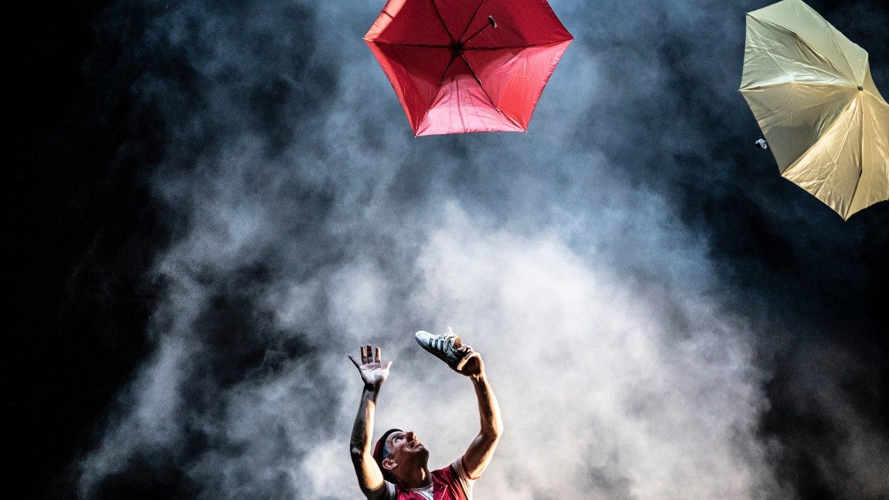  A cast member of Acrobuffos, a white man wearing a red shirt and holding a white Adidas shoe with black stripes in his left hand, both arms raised above his head, gazes upward at two umbrellas, one red and one yellow, floating in the air above him. He's surrounded by smoky fog.
