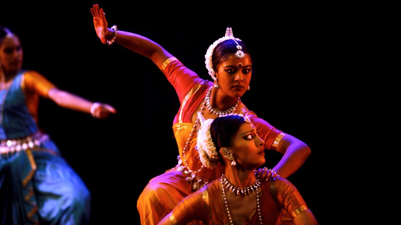 Dancers from "Āhuti" perform on stage in front of a black background. One woman is crouched in front of another, her eyes wide, as the second woman raises her right arm high, as if to strike.