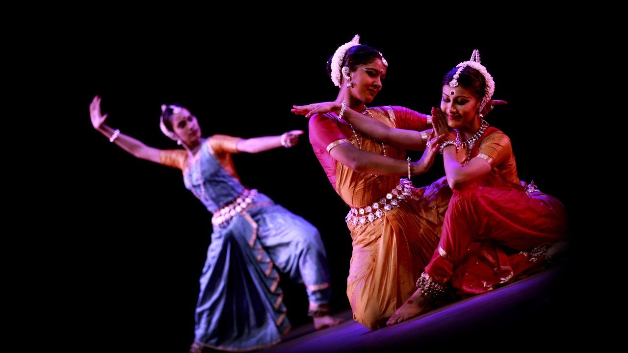  Dancers from "Āhuti" perform on stage in front of a black background. Two women in the foreground touch each other gently and smile, both wearing traditional costumes of orange and red, while a third woman in the background raises her right arm high above and behind her, her left arm extended out in front of her. She wears a traditional costume of blue and orange.