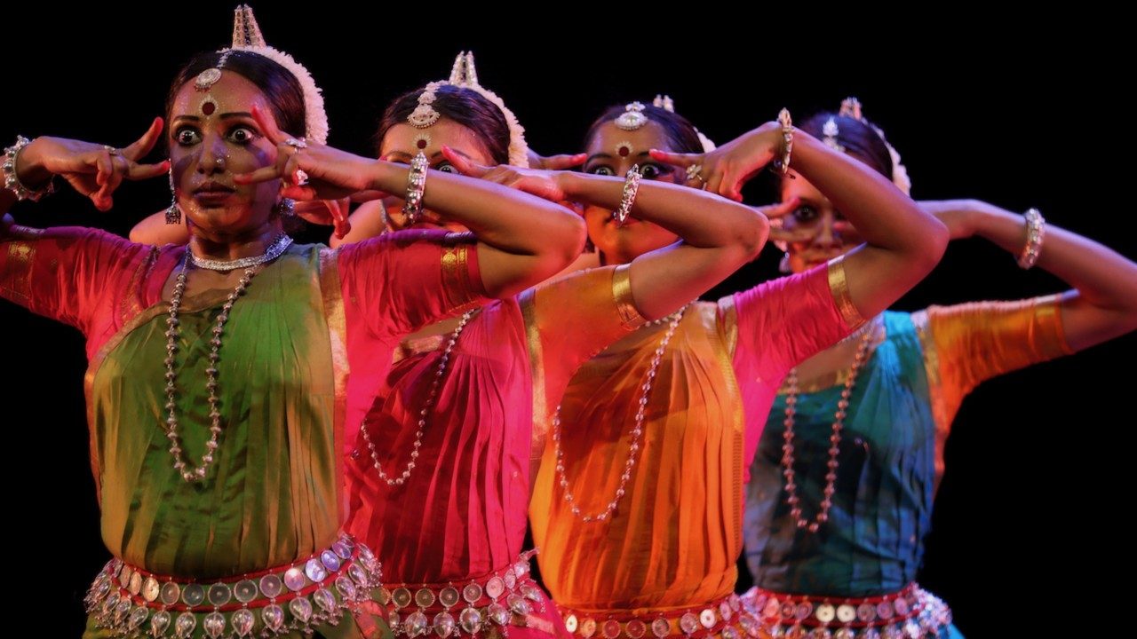 Dancers from "Āhuti" perform on stage in front of a black background. Four women stand in a line, all with both hands towards their eyes, which are open really wide. They wear traditional costumes in rich jewel tones.