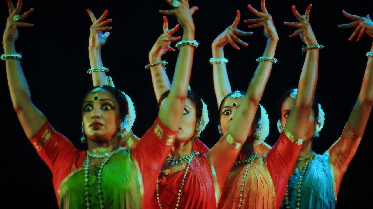  Dancers in "Āhuti" strike poses on stage in front of a black background. Four women raise both arms high above their heads with eyes very wide. They wear traditional costumes in rich jewel tones.
