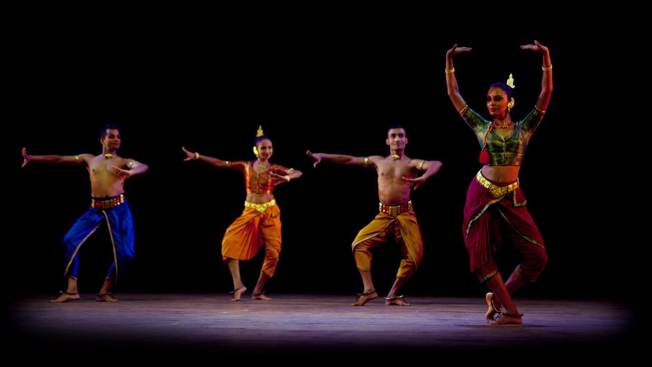 Dancers from "Āhuti" perform on stage in front of a black background. Three line up together in the background, while one woman is in the foreground, both arms raised high above her head. All wear traditional costumes.