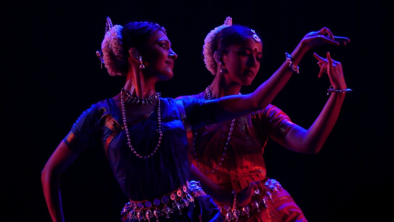  Dancers from "Āhuti" perform on stage in front of a black background. They are lit with blue light and both wear traditional costumes.