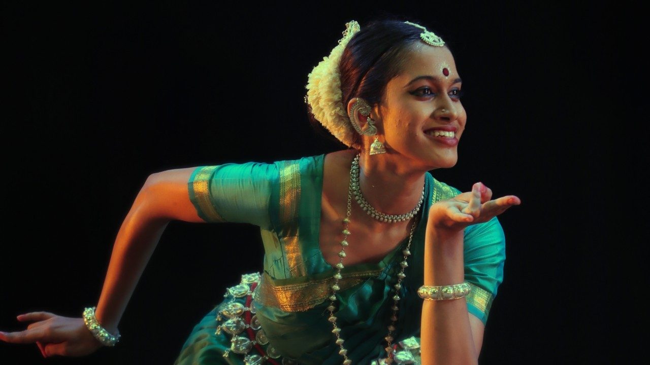  A dancer from "Āhuti" performs on stage in front of a black background, wearing a traditional costume in rich jewel tones.