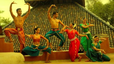  Dancers in "Āhuti" strike poses on a rooftop in front of a clay shingled roof. They wear traditional costumes in oranges, blues, teals, greens, and reds.
