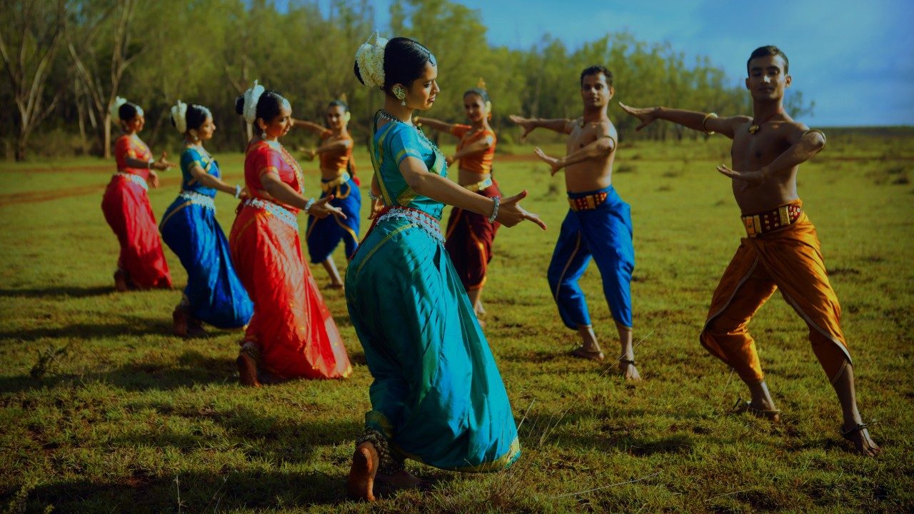  Dancers in "Āhuti" strike poses in a grassy field. They wear traditional costumes in oranges, blues, teals, greens, and reds. They face each other in two lines of four dancers.
