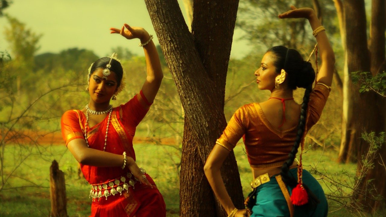  Dancers in "Āhuti" strike poses in a grassy field near a few trees. They wear traditional costumes in oranges, teals, and reds.