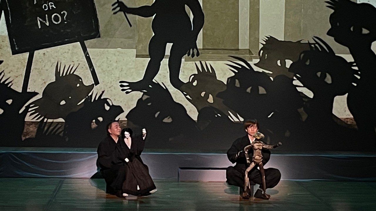  Two men in black operate large puppets. On the wall behind them is a projection of a creature standing up in front of a group of other similar creatures. The standing creature holds a long stick and points to an easel that reads "Does God exist? Yes or no?"