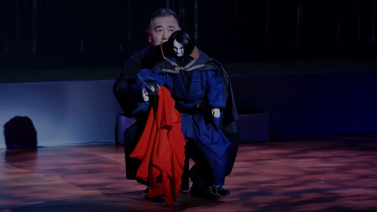  A Japanese man in black operates a large puppet with black hair, blue clothes, and black cloak holding a large red scarf.