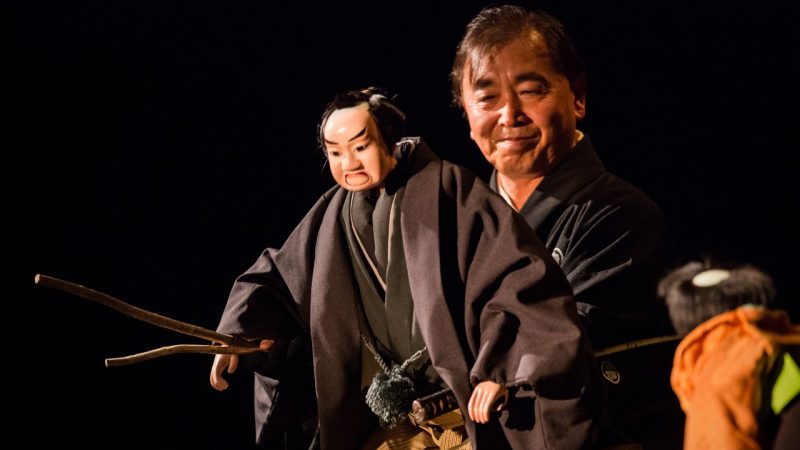  Puppeteer Kory Nishikawa V performs with a Bunraku puppet on stage wearing all back in front of a black background.