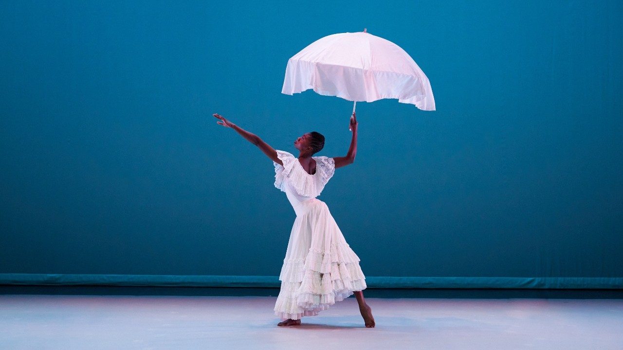 Alvin Ailey American Dance Theater's Khalia Campbell, a Black woman with short braided hair wearing a long white lace dress with tiered ruffles at the hem and one large ruffle around the neckline. She holds a white fabric umbrella overhead, left leg extended with just the toes touching the ground, and her left arm reaching towards the top left corner of the image, her upper body twisted to her right. She stands in front of a deep cerulean background.