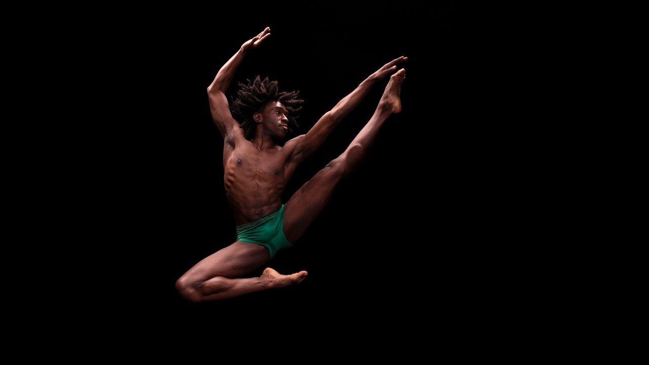 Alvin Ailey American Dance Theater's Chalvar Monteiro, a Black man with medium length brown locs wearing emerald green shorts, jumps into the air, his right leg bent at the knee underneath him, left leg fully extended and his left hand reaching past his left toes. His right arm is bent up overhead, palm facing away from his body. He jumps in front of a black background.