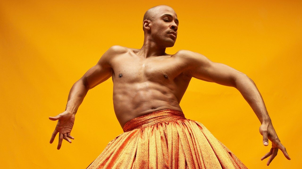Alvin Ailey American Dance Theater's Yannick Lebrun, photo by Dario Calmese. Lebrun, a Black man with a shaved head, wears a long, full, glittery orange skirt and no shirt. He  holds his arms out at angles on either side of his body in front of an orange background.