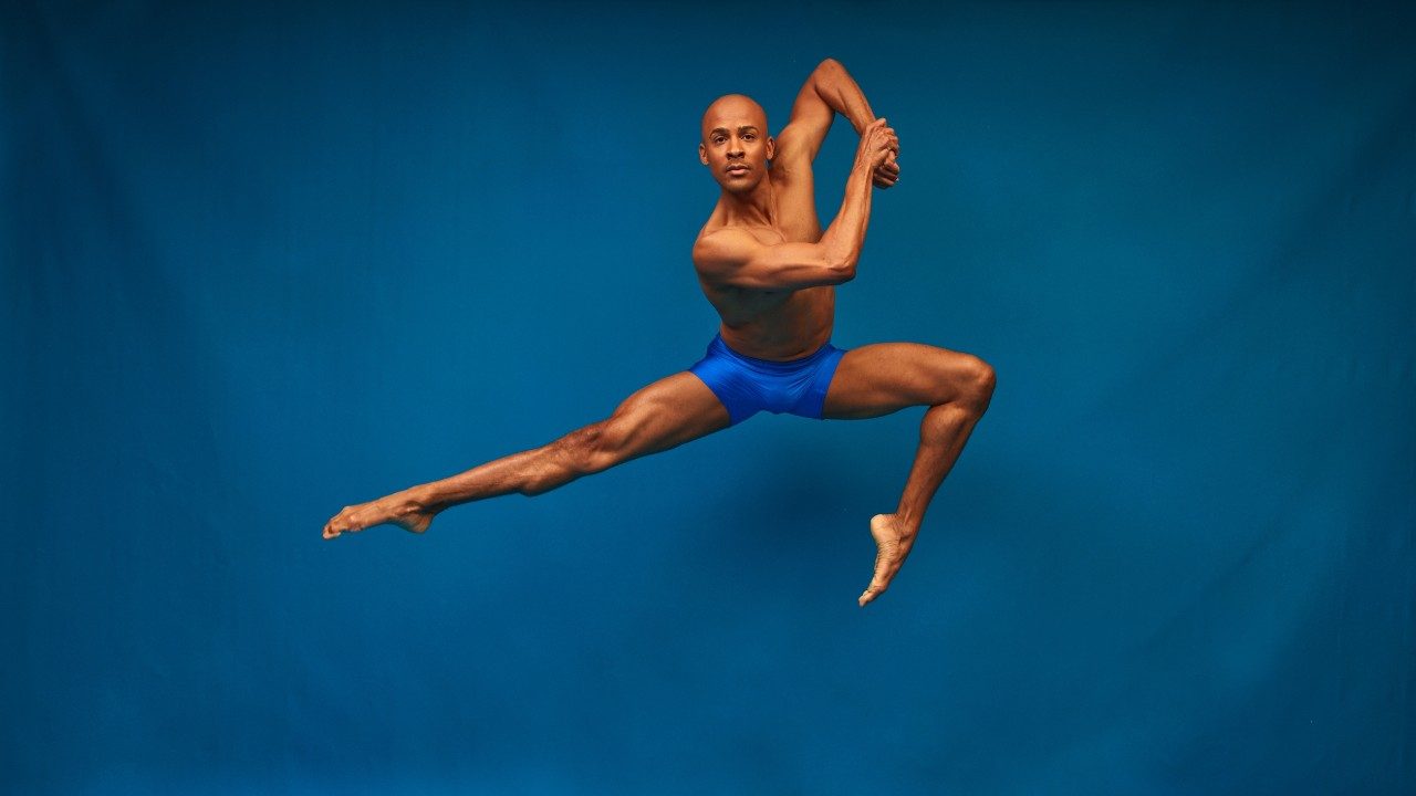  Alvin Ailey American Dance Theater's Yannick Lebrun, photo by Dario Calmese. Leburn, a light skinned bald Black man, wears blue shorts and jumps into the air in front of a blue background.