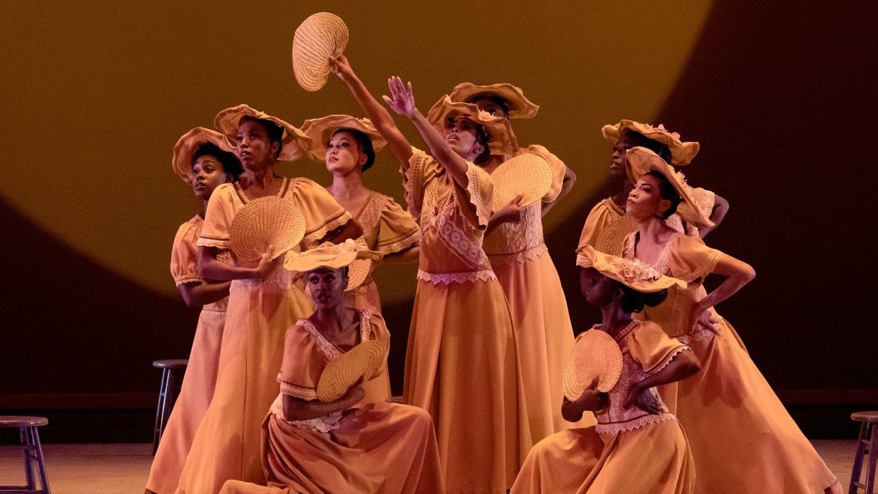  Alvin Ailey American Dance Theater performs "Revelations," photo by Paul Kolnik. A group of nine Black women in long tan dresses and hats holding rattan hand fans perform on stage.