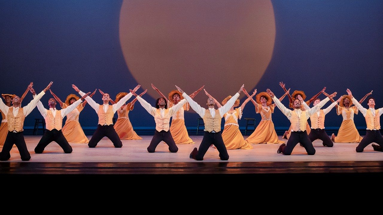  Alvin Ailey American Dance Theater performs "Revelations," photo by Paul Kolnik. Eight men in black pants, white shirts, and tan vests, and eight Black women in long tan dresses and tan hats, all kneel on stage, both arms extended overhead in a V shape. Behind them is a large projection of a moon-like object against a navy blue background.