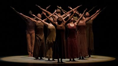  Alvin Ailey American Dance Theater performs "Revelations" wearing monochrome costumes in various shades of brown. They all stand with both arms straight and in a wide V overhead.