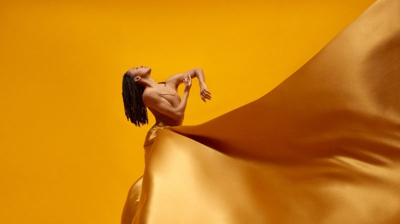 Alvin Ailey American Dance Theater's Jacquelin Harris, photo by Dario Calmese. Harris, a light skinned Black woman with medium length braids, is draped in a gold dress, her face tilted towards the sky. The background is a similar gold tone to her dress.