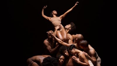  Members of Alvin Ailey American Dance Theatre wear skin-toned body suits and shorts. They gather crouched together against a black background, a Black woman with natural hair is lifted above the rest by two Black men, her arms extended gracefully.