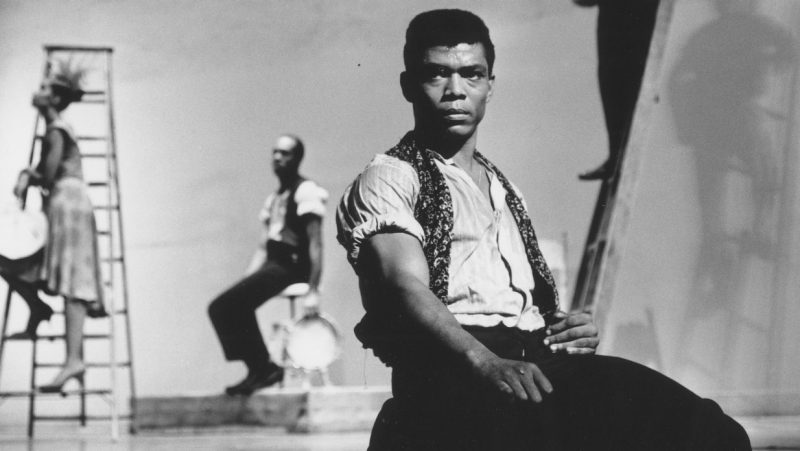 A black and white still from "American Masters: Ailey," which shows choreographer and dancer Alvin Ailey kneeling on stage in the foreground.