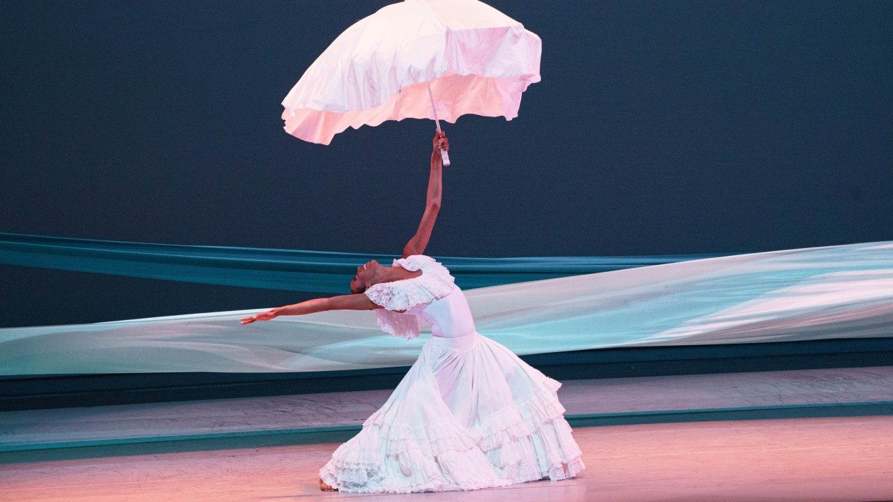  Alvin Ailey American Dance Theater's Courtney Celeste Spears, photo by Daniel Azoulay. Spears, a Black woman in a long ruffled white dress, leans one arm and her head backwards and holds an umbrella with draped silk fabric above her head while performing "Revelations" on stage. Behind her is a navy blue background and two long pieces of fabric, one teal and one white, that stretch from side to side across the stage.