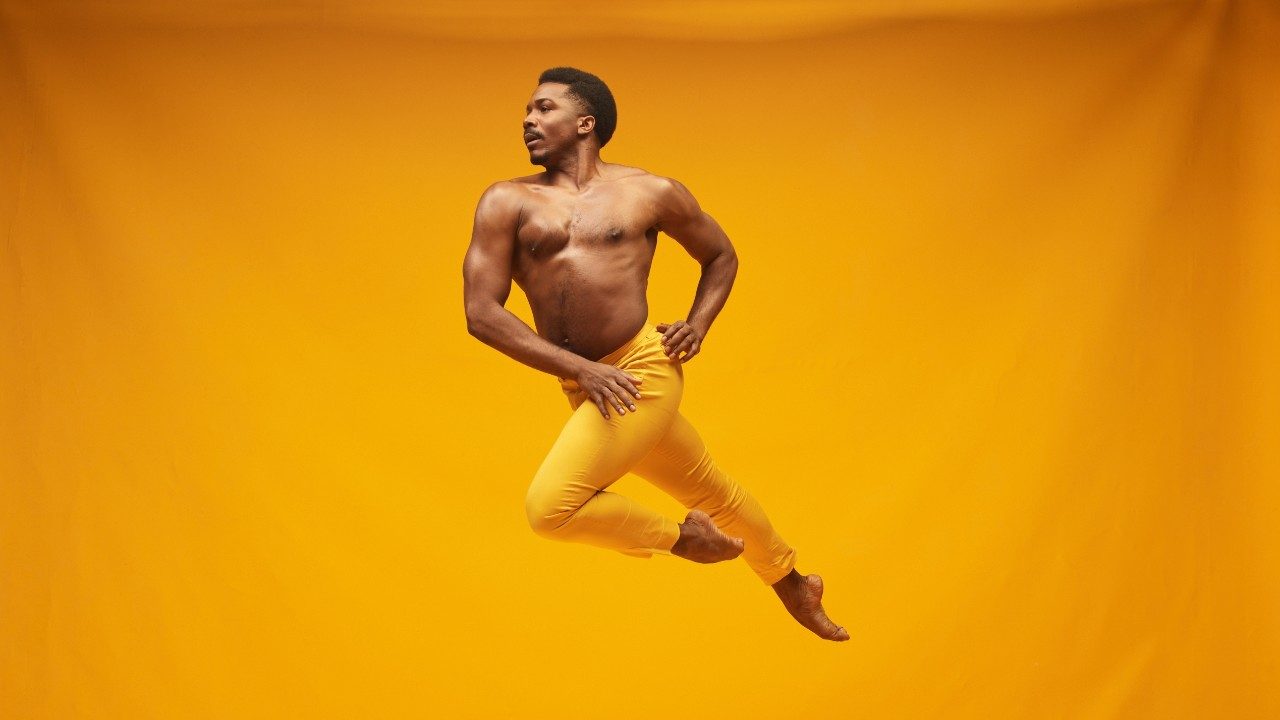 Alvin Ailey American Dance Theater's Michael Jackson Jr., photo by Dario Calmese. Jackson is a Black man with a short Afro. He wears gold pants and jumps into the air in front of a matching gold background.