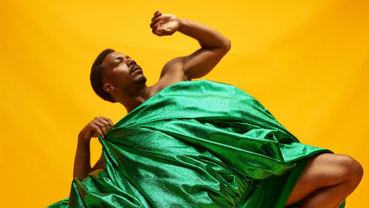  Alvin Ailey American Dance Theater's Michael Jackson Jr., photo by Dario Calmese. Jackson is a Black man with short Afro. He wears a long sparkly green skirt and kneels on the ground in front of a gold background.