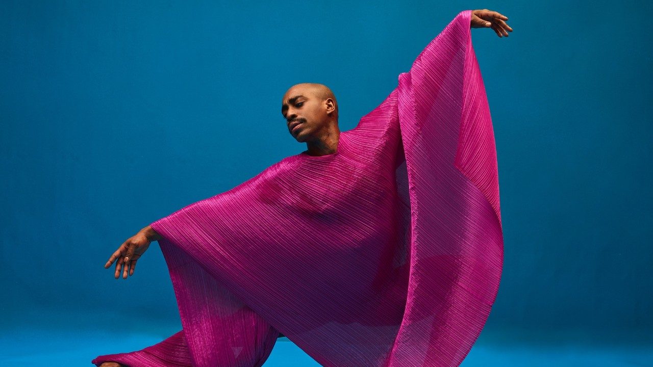  Alvin Ailey American Dance Theater's Renado Maurice, photo by Dario Calmese. Maurice, a light skinned bald Black man, wears a flowing magenta poncho and extends both arms gracefully in front of a navy blue background.