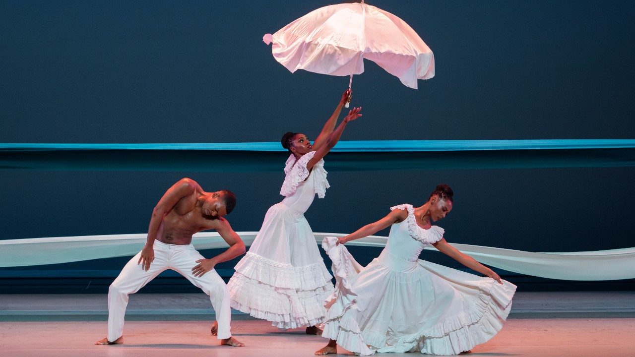  Alvin Ailey American Dance Theater's Solomon Dumas, Khalia Campbell, and Samantha Figgins in "Revelations," photo by Paul Kolnik. The dancers, two Black women in long pale ruffled dresses and one Black man in white pants, dance on stage. The woman in the middle holds an umbrella draped in silk fabric. Behind them, two large scraps of fabric extend across the width of the stage in front of a navy blue background.