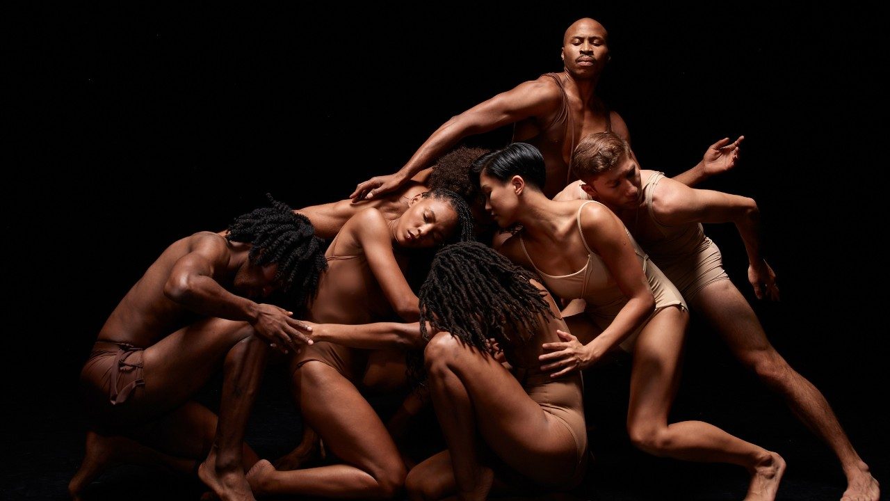  Members of Alvin Ailey American Dance Theatre wear skin-toned body suits and shorts. They gather crouched together against a black background, a Black man with a bald head and a moustache rises up above the others.