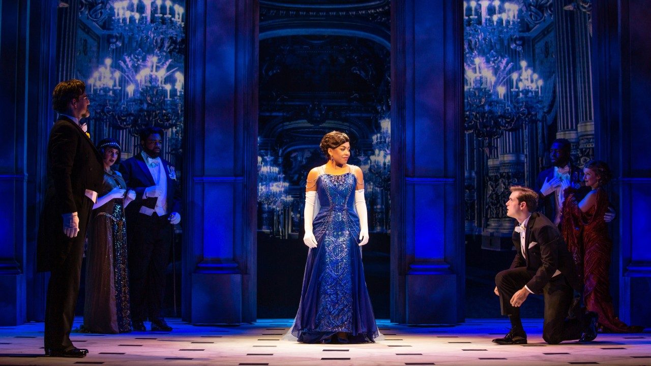  A light skinned Black woman wears a floor length royal blue dress and long white gloves. She stands in front of an ornate hall in a palace and gazes towards a white man, who kneels in a tuxedo as if fixing his shoe. Other couples stand nearby and watch the pair.