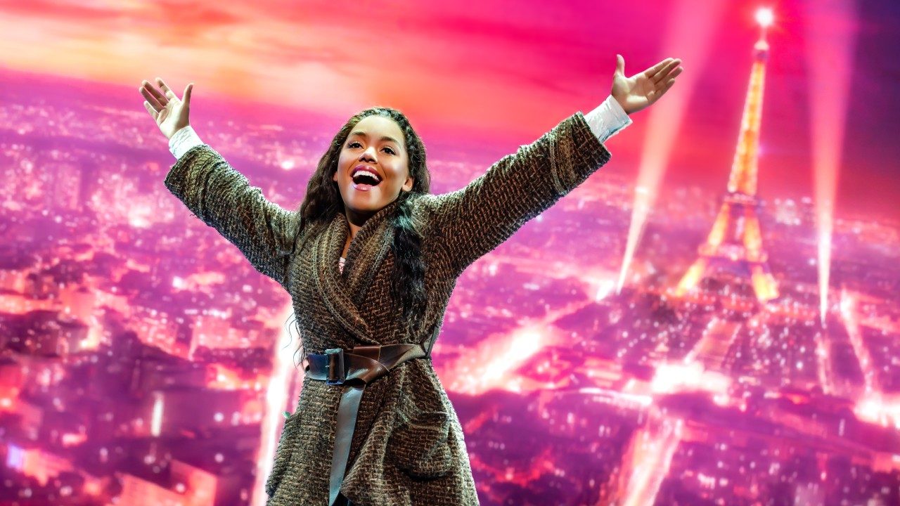  Anastasia, played by Kyla Stone, a light-skinned Black woman with long brown hair, stands in front of a pink background image of Paris, the Eiffel Tower prominently visible. She wears a long, tweed, brown dress belted at the waist, both arms outstretched wide.