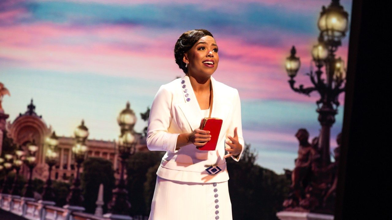  A light skinned Black woman wears a white skirt suit set, her brown hair pulled into a sleek updo. She holds a small red pocketbook and behind her are Parisian street lights, a bridge, and a large columned building at sunset.