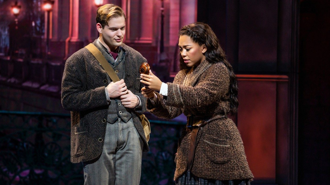  A white man in clothes from the early 1900s stands next to a light skinned Black woman in a long brown tweed dress. She holds a music box and looks wonderingly at it.