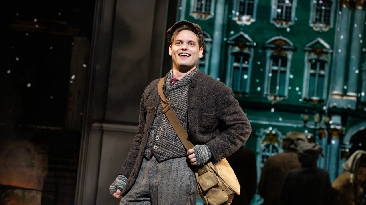  A white man wears 1920s-era clothes: a brown jacket, brown striped pants, brown vest, grey gloves, and a tan messenger bag. He smiles towards the audience.
