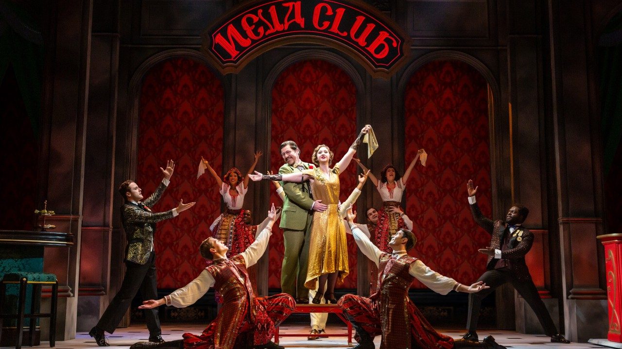  The cast of "Anastasia" poses during a musical number. They wear clothes from the early 1900s, two people coupled together in the center of the others. Behind them is red patterned wallpaper and large intricate wood paneling. A red sign above them reads "Neva Club"