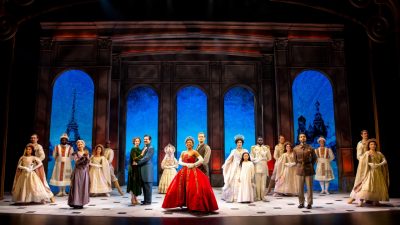  The cast of "Anastasia" poses onstage, most people are in pairs. In the center is a light skinned Black woman wearing a long red ball gown and crown and a white man in darks pants and a vest.