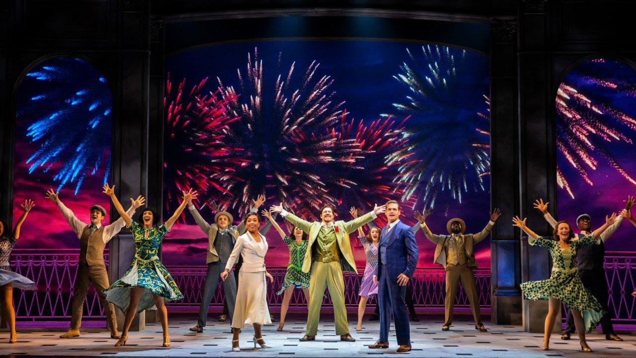  The cast of "Anastasia" sings and dances on stage, projections of fireworks visible behind them. Nearly everyone stands with their arms raised above their heads, outstretched wide.